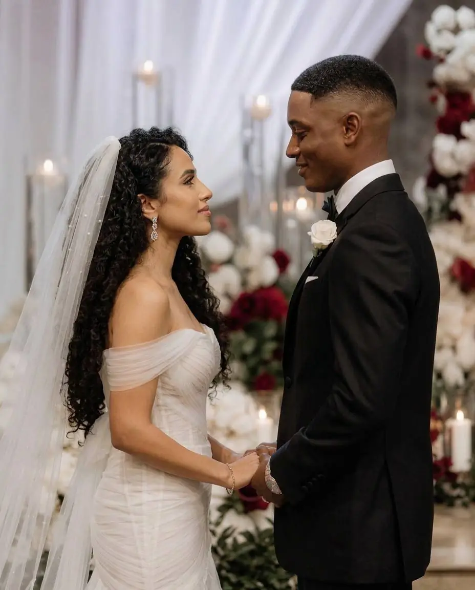 A bride in a veil and her groom holding hands at a candlelit altar surrounded by white and deep red roses and white drapery